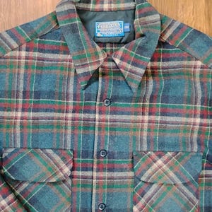 May include: A long-sleeve button-up shirt featuring a plaid pattern in shades of teal, dark blue, red, and tan. The shirt includes two chest pockets with flaps, a pointed collar, and a blue Pendleton brand label sewn inside the neck.