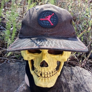 K&ouml;nnte beinhalten: Eine braune Baseballkappe mit einem schwarzen und roten Logo, das eine springende Figur in einem Kreis zeigt. Die Kappe hat einen schwarz-wei&szlig;en gemusterten Schirm.