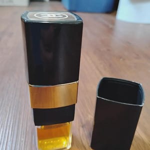 May include: A rectangular perfume bottle with a black cap and a gold-toned band around the middle. The top of the cap features a white Chanel logo. The glass base of the bottle contains a golden-yellow liquid, and a separate black cap sits nearby.