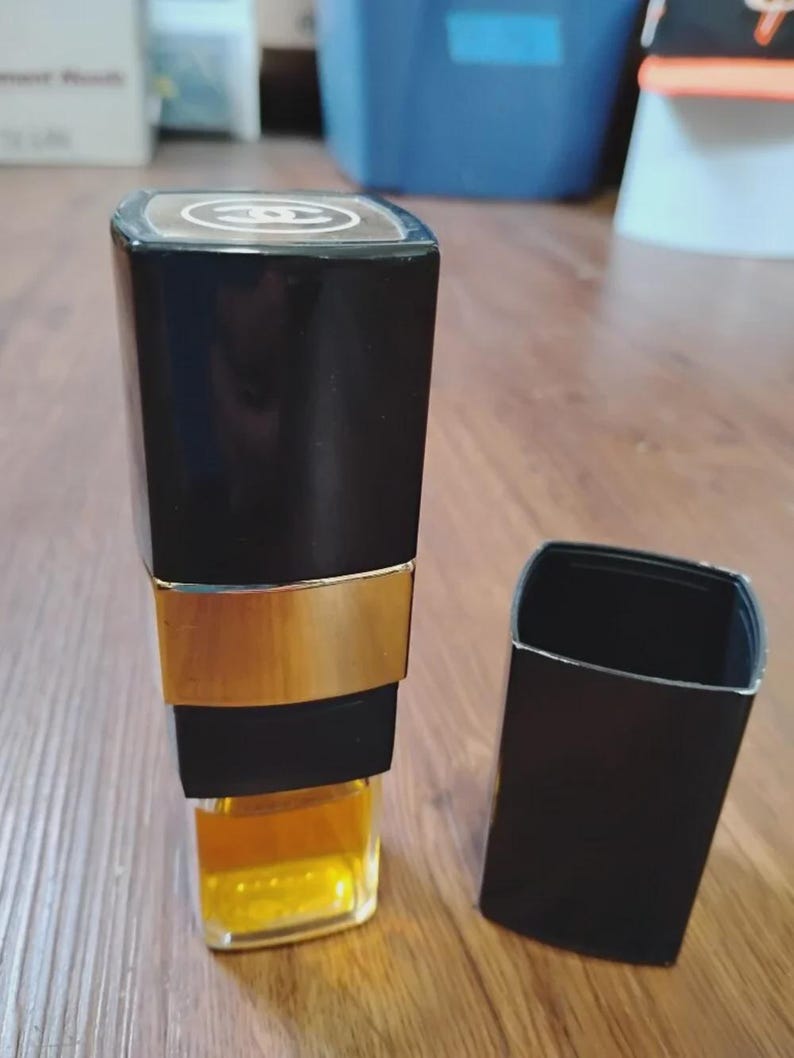 May include: A rectangular perfume bottle with a black cap and a gold-toned band around the middle. The top of the cap features a white Chanel logo. The glass base of the bottle contains a golden-yellow liquid, and a separate black cap sits nearby.