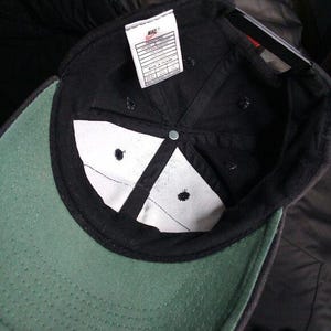 May include: Black baseball cap with a green brim and white interior panels. The cap features a Nike tag and a metal button at the crown. The interior has a white lining and black stitching.