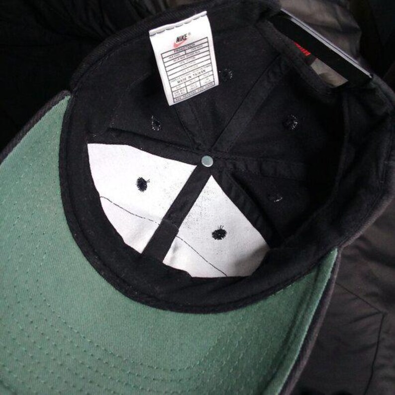 May include: Black baseball cap with a green brim and white interior panels. The cap features a Nike tag and a metal button at the crown. The interior has a white lining and black stitching.