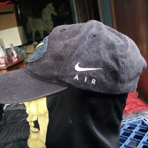 May include: A black baseball cap with a curved brim. The Nike logo and the word "AIR" are embroidered in white on the side. A patch is visible on the side of the cap.