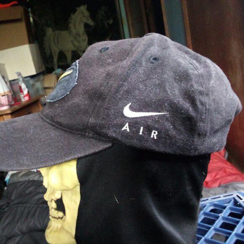 May include: A black baseball cap with a curved brim. The Nike logo and the word "AIR" are embroidered in white on the side. A patch is visible on the side of the cap.