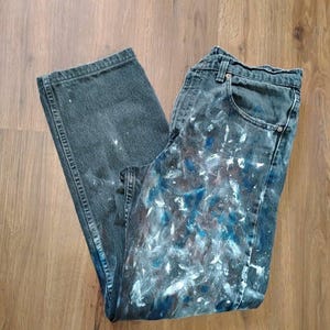 Puede incluir: Unos vaqueros de mezclilla gris oscuro con un diseño de salpicaduras de pintura en la parte delantera. La tela tiene una textura acanalada vertical sutil y está decorada con manchas irregulares de pintura blanca, azul y marrón por toda la prenda.
