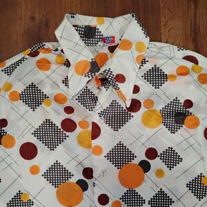 Könnte beinhalten: Ein Vintage-Hemd mit Knöpfen und weißem Hintergrund, das ein sich wiederholendes Muster aus orangefarbenen, braunen und schwarzen Kreisen und karierten Quadraten aufweist. Das Hemd hat einen spitzen Kragen und einen Knopfverschluss. Das Design des Hemdes erinnert an die 1970er Jahre.