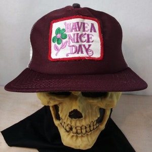 Op de afbeelding: Een bordeauxrode truckercap met een wit mesh achterpaneel. De voorkant van de cap heeft een rood, wit en groen geborduurd patch met de tekst "Have a Nice Day" met een bloemdessin.