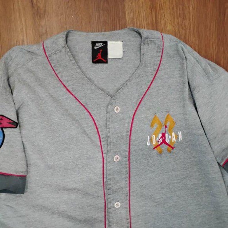 May include: A grey baseball jersey with red trim and a button-up front. The jersey features the Nike logo and a "23 Jordan" emblem on the chest. The sleeves have red and grey cuffs.