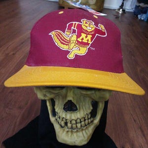 Puede incluir: Gorra de béisbol granate y dorada con una mascota de ardilla de dibujos animados con la letra "M". La gorra tiene una visera plana y una corona estructurada. La ardilla lleva una camiseta con la letra "M".