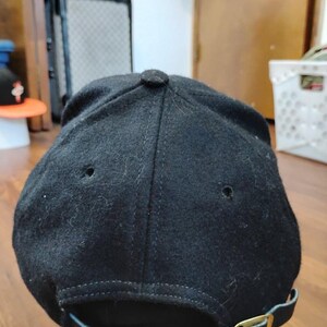 May include: A black baseball cap with a felt-like texture, featuring a six-panel construction with stitched eyelets on the crown. The back includes an adjustable black strap with a metallic gold-toned buckle for a secure fit and closure.