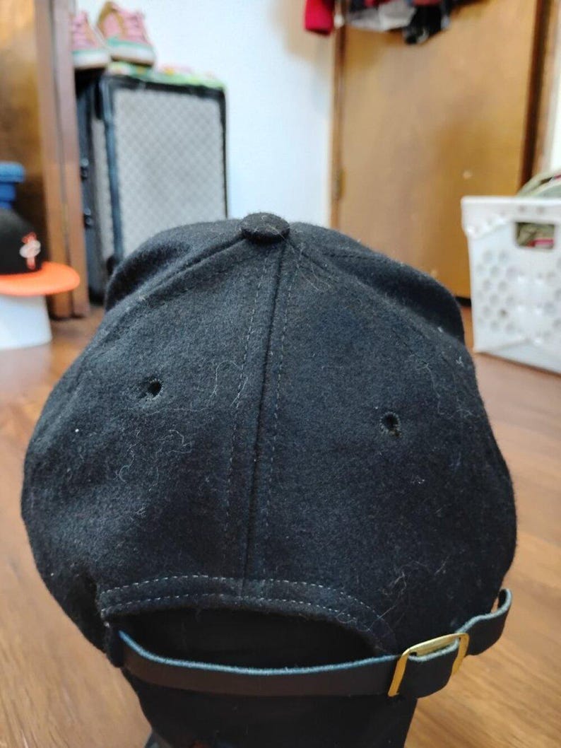 May include: A black baseball cap with a felt-like texture, featuring a six-panel construction with stitched eyelets on the crown. The back includes an adjustable black strap with a metallic gold-toned buckle for a secure fit and closure.