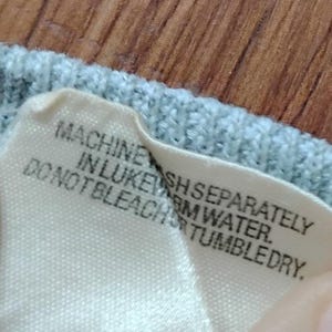 May include: Close-up of a light blue knit fabric with a white tag attached. The tag has black text that reads: "MACHINE WASH SEPARATELY IN LUKEWARM WATER. DO NOT BLEACH OR TUMBLE DRY."