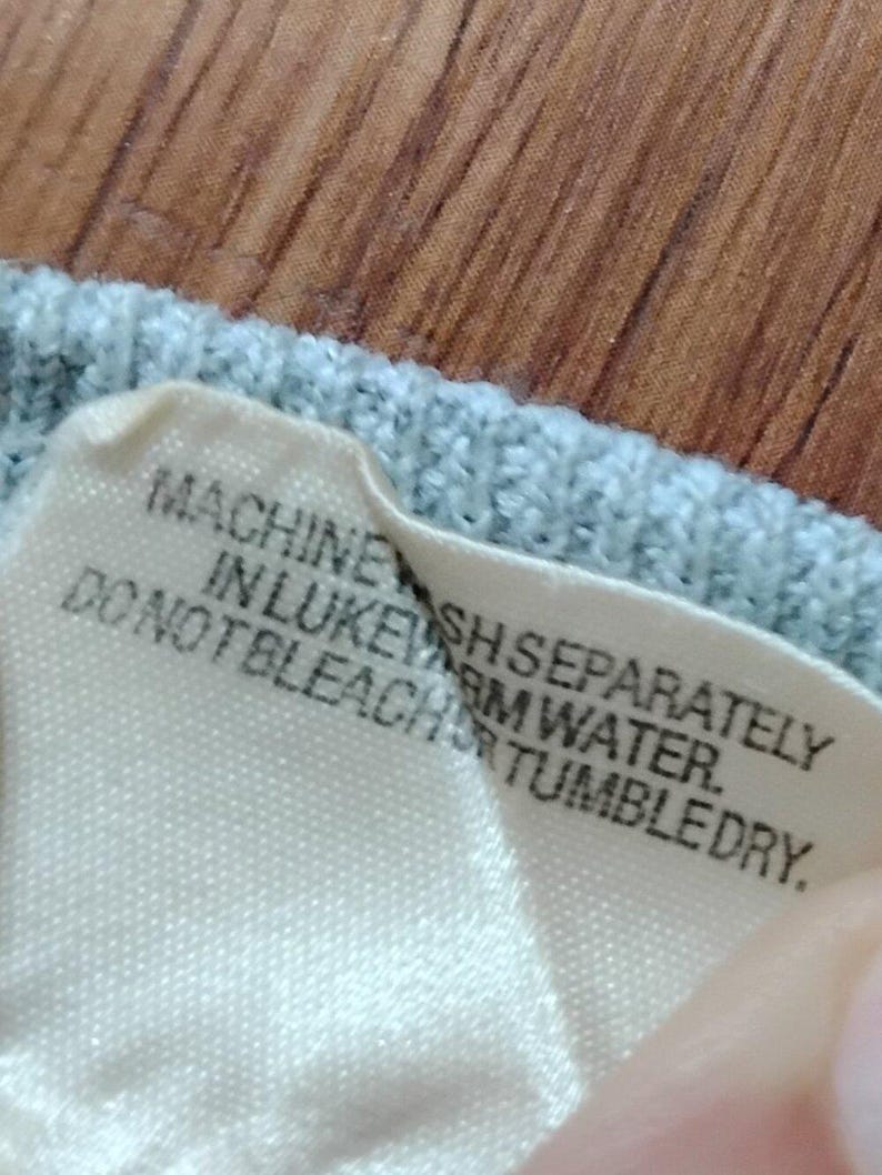 May include: Close-up of a light blue knit fabric with a white tag attached. The tag has black text that reads: "MACHINE WASH SEPARATELY IN LUKEWARM WATER. DO NOT BLEACH OR TUMBLE DRY."