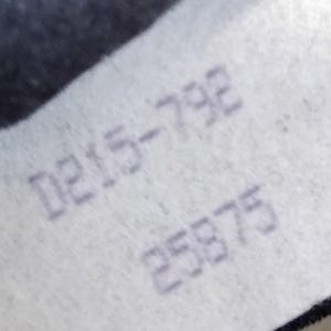 May include: A white fabric tag featuring the printed text D215-792 and 25875 in a dark gray font. The tag has a slightly textured surface and is positioned against a dark, out-of-focus background, displaying clear alphanumeric characters.