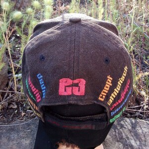 K&ouml;nnte beinhalten: Eine braune Baseballkappe mit schwarzem Schirm und einem roten und wei&szlig;en Logo mit der Aufschrift "GO". Die Kappe hat einen bunten gestickten Text um den Schirm herum, darunter "Uni&atilde;o", "Coupi" und "millenrico".