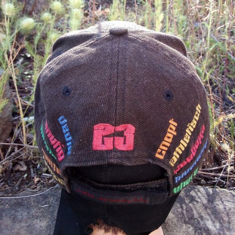 K&ouml;nnte beinhalten: Eine braune Baseballkappe mit schwarzem Schirm und einem roten und wei&szlig;en Logo mit der Aufschrift "GO". Die Kappe hat einen bunten gestickten Text um den Schirm herum, darunter "Uni&atilde;o", "Coupi" und "millenrico".