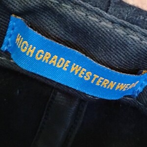 May include: A black fabric cap featuring an interior blue rectangular tag stitched onto the inner band. The tag displays the yellow text HIGH GRADE WESTERN WEAR in a bold, sans-serif font, clearly visible against the dark material of the cap.