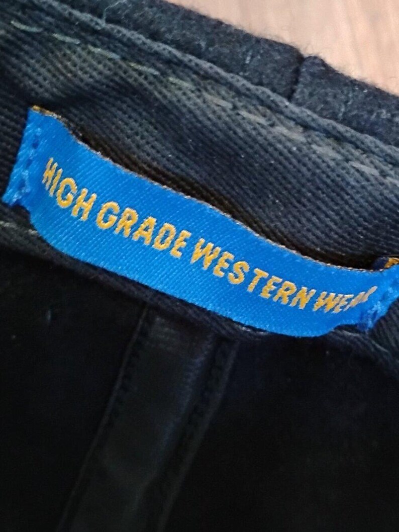 May include: A black fabric cap featuring an interior blue rectangular tag stitched onto the inner band. The tag displays the yellow text HIGH GRADE WESTERN WEAR in a bold, sans-serif font, clearly visible against the dark material of the cap.