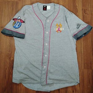 May include: Grey baseball jersey with red trim and a button-up front. It features the "Jordan" logo on the chest and sleeve patches, along with Nike logos. The jersey has short sleeves with dark grey cuffs.