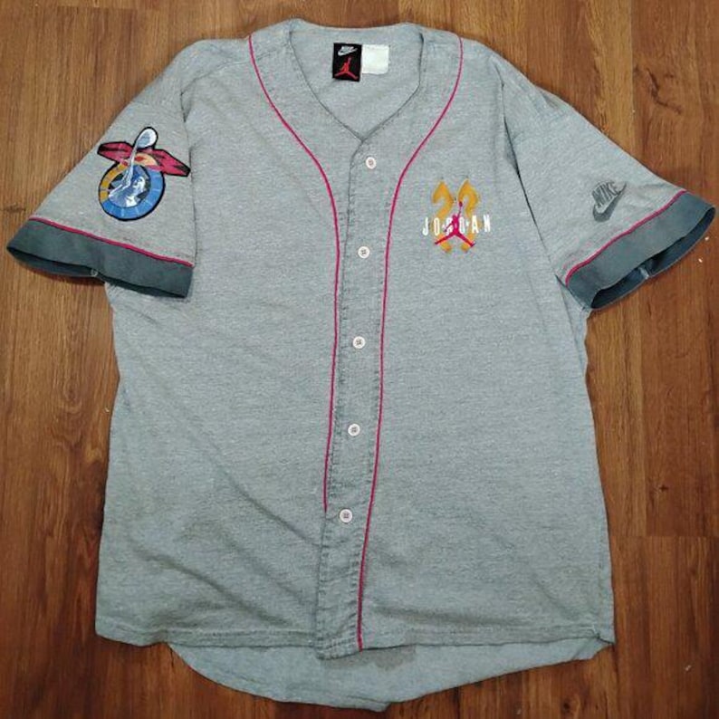 May include: Grey baseball jersey with red trim and a button-up front. It features the "Jordan" logo on the chest and sleeve patches, along with Nike logos. The jersey has short sleeves with dark grey cuffs.