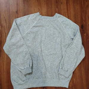 May include: A heather gray sweatshirt with long sleeves and a crew neck. The sweatshirt has a relaxed fit and a simple, classic design. The sleeves are raglan style, and the cuffs and hem are ribbed.