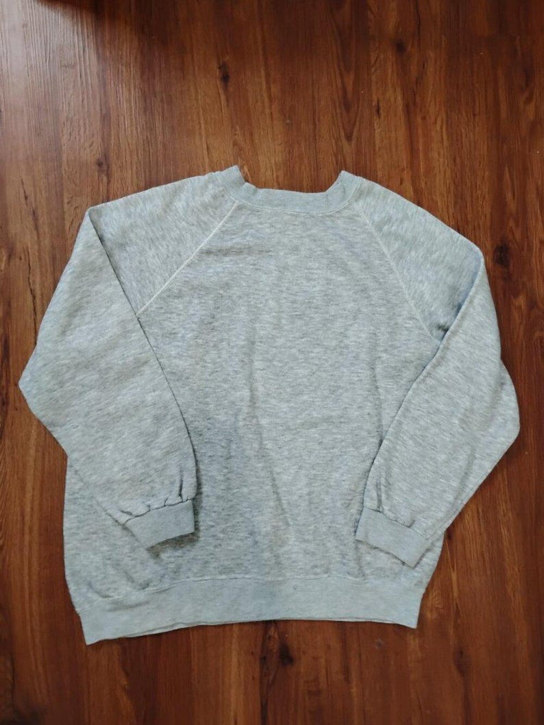 May include: A heather gray sweatshirt with long sleeves and a crew neck. The sweatshirt has a relaxed fit and a simple, classic design. The sleeves are raglan style, and the cuffs and hem are ribbed.