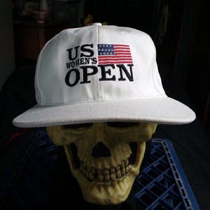 Op de afbeelding: Witte baseballcap met de tekst "US Women's Open" en een Amerikaanse vlag. De pet wordt gedragen door een schedel.