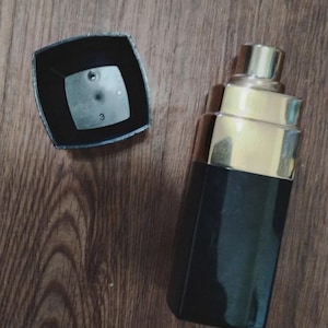 May include: A rectangular perfume bottle with a black base and a gold-toned stepped neck. The matching black square cap is detached and placed beside the bottle, revealing the number 3 printed on the interior surface of the cap's top panel.