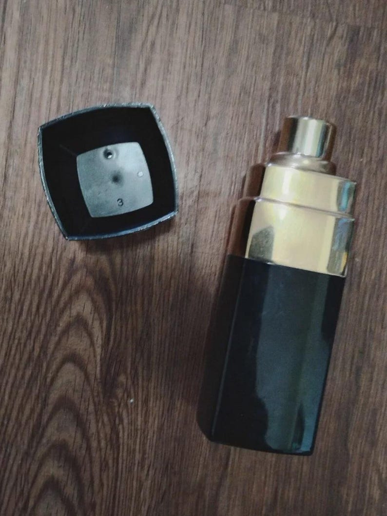May include: A rectangular perfume bottle with a black base and a gold-toned stepped neck. The matching black square cap is detached and placed beside the bottle, revealing the number 3 printed on the interior surface of the cap's top panel.