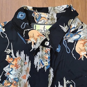 Pode incluir: Camisa preta com padrão colorido apresentando figuras, cavalos e elementos florais. A camisa tem gola clássica e punhos abotoados. A etiqueta da marca "Loco Live" é visível.