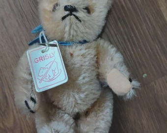 Puede incluir: Un oso de peluche beige con una cinta azul alrededor del cuello y una etiqueta blanca unida a la cinta. La etiqueta presenta un borde rojo, el texto GRISLY y un dibujo lineal de un oso. El oso tiene ojos oscuros, una nariz cosida y patas.