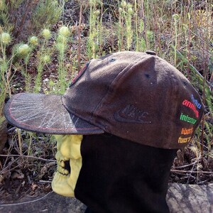 K&ouml;nnte beinhalten: Eine braune Baseballkappe aus Cord mit rotem Schirm und einem Nike-Logo an der Seite. Die Kappe hat einen bunten gestickten Text an der Seite, der "indie amazing interior lovers forever" lautet.