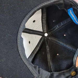 May include: The interior of a black baseball cap featuring a white fabric lining, black structural seams, and a small blue tag with the text HIGH GRADE WESTERN. A single metallic button is visible at the center crown and a brass snap closure at the back.