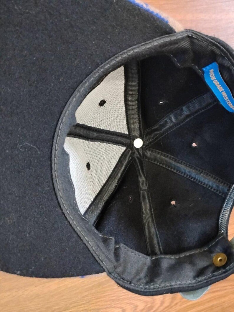 May include: The interior of a black baseball cap featuring a white fabric lining, black structural seams, and a small blue tag with the text HIGH GRADE WESTERN. A single metallic button is visible at the center crown and a brass snap closure at the back.