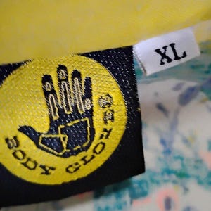 Puede incluir: Primer plano de una etiqueta negra y amarilla con las palabras "BODY GLOVE" y un logotipo de una mano. La etiqueta tambi&eacute;n tiene una etiqueta blanca con "XL" impresa. La etiqueta est&aacute; adherida a una prenda amarilla.