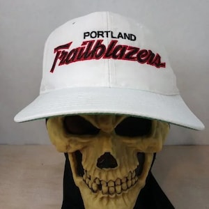 Może przedstawiać: Biała czapka bejsbolówka z haftowanym logo w kolorze czarnym i czerwonym, na którym widnieje napis "PORTLAND Trailblazers".