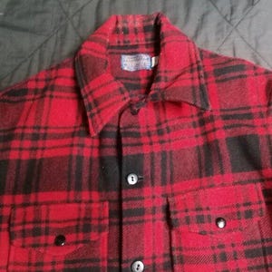 May include: A red and black plaid button-up shirt with two chest pockets and a collar. The shirt has black buttons and a small label on the collar. The fabric appears to be a thick, woven material.