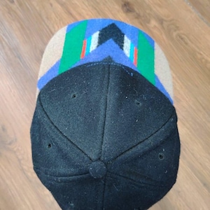 May include: A baseball cap featuring a solid black crown and a brim with a geometric pattern. The brim displays a tan base with blue, green, white, and red angular stripes arranged in a chevron-like design. The cap has six panels and a button.