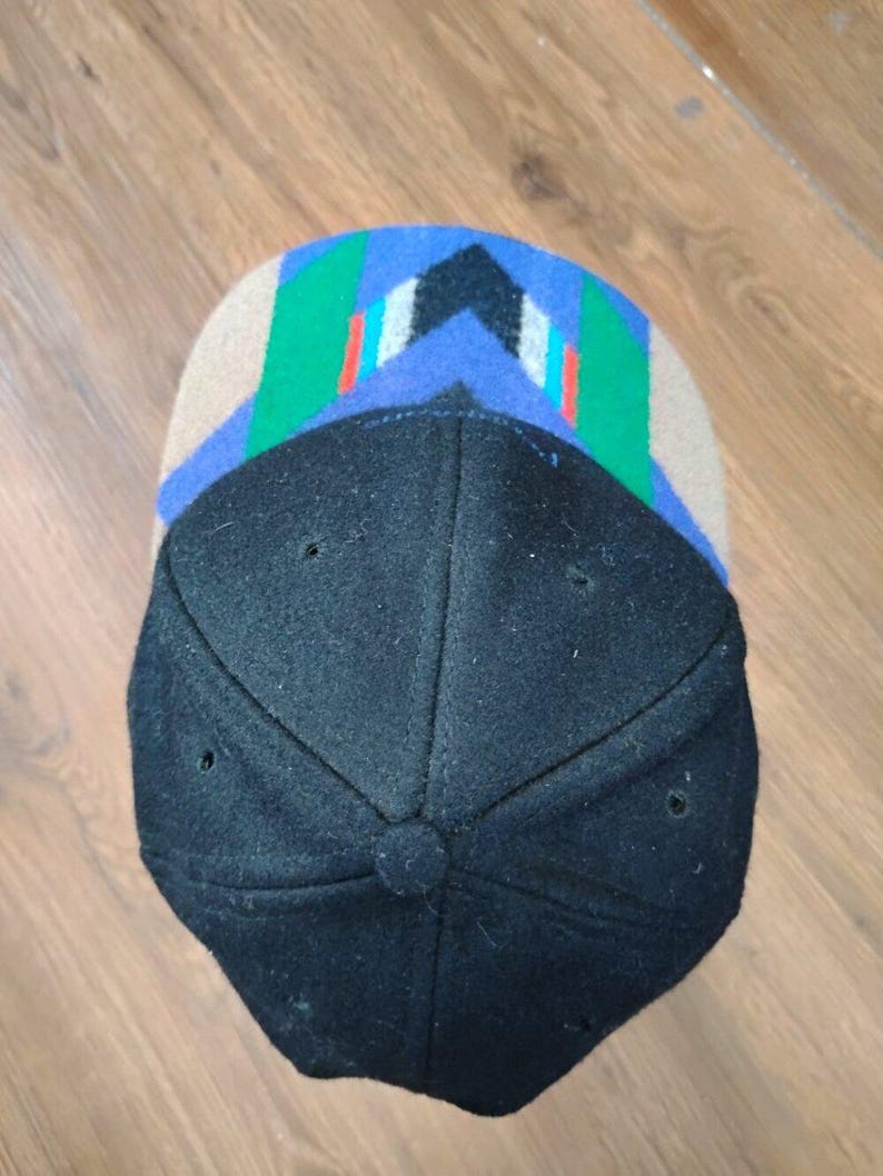 May include: A baseball cap featuring a solid black crown and a brim with a geometric pattern. The brim displays a tan base with blue, green, white, and red angular stripes arranged in a chevron-like design. The cap has six panels and a button.