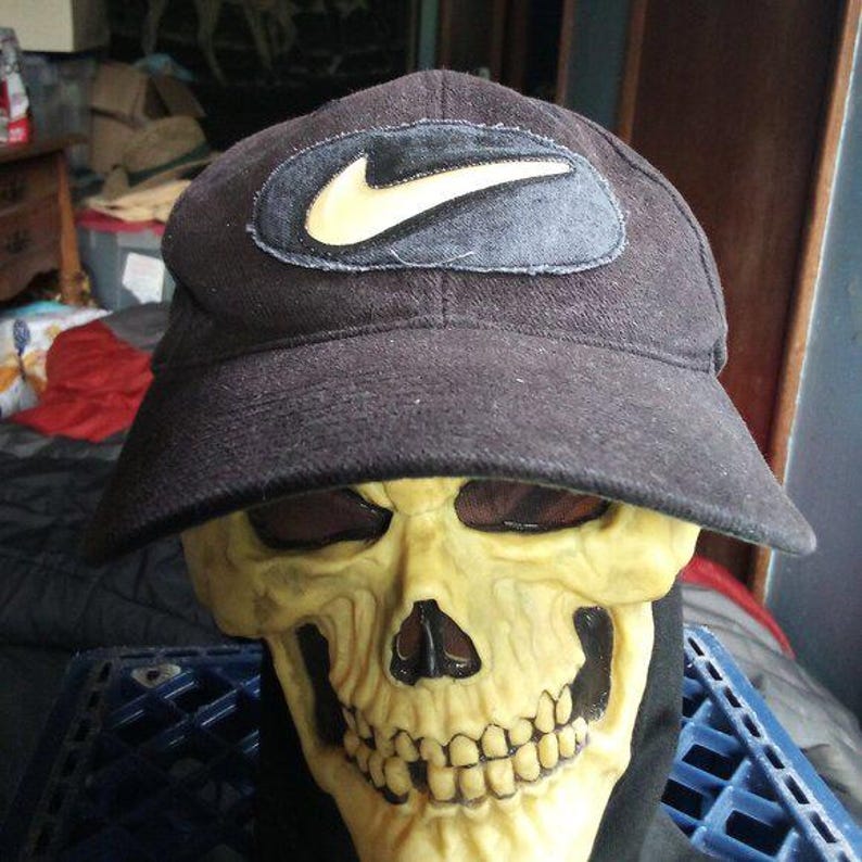 May include: A black baseball cap with a yellow Nike logo patch is worn by a skull mask. The skull mask is yellow with dark eye sockets and a wide grin. The cap's brim casts a shadow over the mask's eyes.