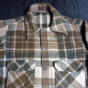 May include: Close-up of a button-down plaid shirt in shades of brown, green, and cream. The shirt features two chest pockets and a classic collar. The fabric appears to be a medium-weight material, suitable for cooler weather.