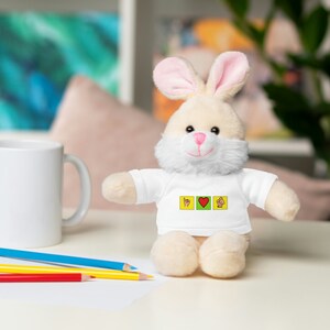 Op de afbeelding: Een wit pluche konijn dat een wit T-shirt draagt met een geel, rood en groen ontwerp. Het ontwerp bevat drie vierkanten met een handgebaar, een hart en een ander handgebaar.