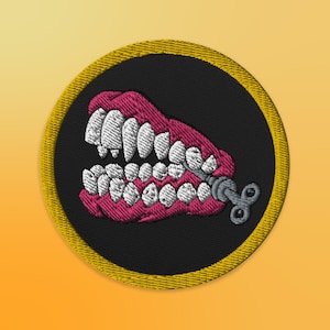 Könnte beinhalten: Ein runder, schwarzer gestickter Aufnäher mit gelbem Rand. Der Aufnäher zeigt ein rosafarbenes und weißes Cartoon-Gebiss mit einem aufgewickelten Schlüssel.
