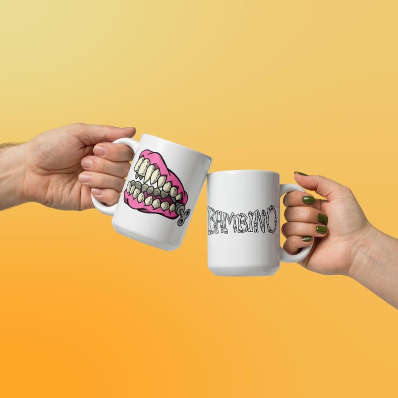 May include: Two white ceramic mugs. One mug has a pink and yellow cartoon image of a set of dentures with a winder key. The other mug has the word 'KAMBINO' written in black letters.