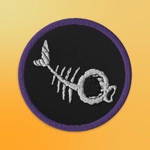 May include: A purple and black embroidered patch featuring a white fish skeleton design. The fish skeleton is stylised with a large open mouth.