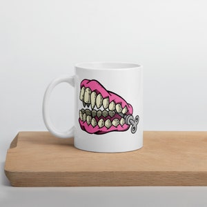 May include: A white coffee mug with a pink and white cartoon image of a set of dentures with a winder key on the side.