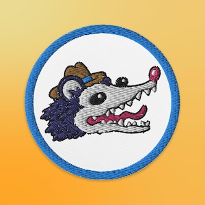 Puede incluir: Parche bordado con un opossum de dibujos animados que lleva un sombrero de vaquero marrón con una banda azul. El opossum tiene una cara blanca con la lengua rosa que sobresale y un pelaje azul y morado.