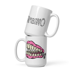 May include: Two white ceramic mugs stacked on top of each other. The top mug has the word 'Symbiotic' written in black on the front. The bottom mug has a pink cartoon image of a set of dentures with a wind-up key on the side.