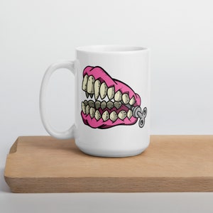 May include: A white ceramic mug with a pink and white cartoon image of a set of dentures with a winder key on the side.