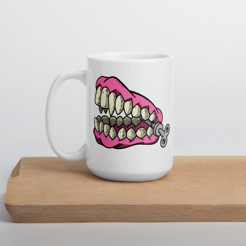 May include: A white ceramic mug with a pink and white cartoon image of a set of dentures with a winder key on the side.